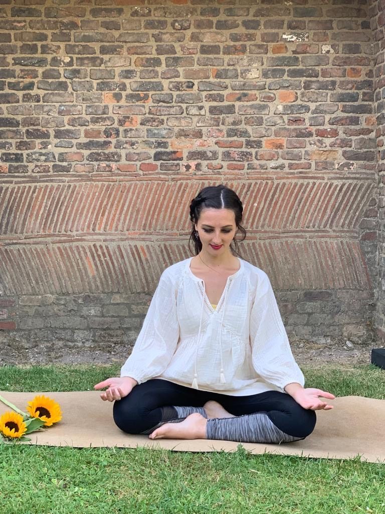 Ria sitting cross-legged in meditation on a mat on the grass, beside sunflowers and an old brick wall